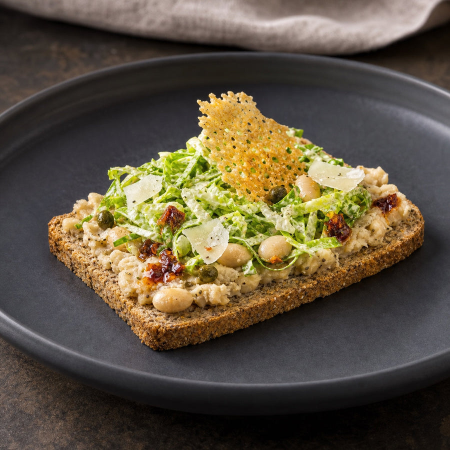 Eine dünne Scheibe Saftkorn Lupine mit Bohnencreme, Caesar-Salat, Parmesan-Crunch und Chiliöl